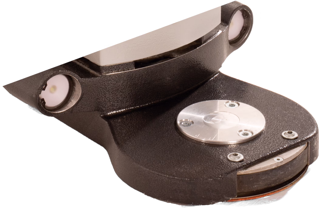 PALLMANN_dummy_Gecko Star attachment length short_msl_2018-07 Nahaufnahme eines kleinen industriellen elektromechanischen Bauteils vor weißem Hintergrund. Das Bauteil zeigt ein strukturiertes, dunkel lackiertes Metallgehäuse mit geschwungenen Konturen, eine polierte, flache Metallkontaktfläche an der Oberseite und eine runde, bearbeitete silberne Scheibe, die mit drei kleinen Schrauben befestigt ist. Nahe den oberen Kanten sind zwei eingelassene, kreisrunde LED-ähnliche Module oder Anzeige-Linsen mit weißen Diffusoren und Metallringen zu sehen. Am unteren Rand erkennt man eine abgerundete Basis mit weiteren Schrauben, eine dünne orangefarbene Dichtung oder Isolationsschicht und einen Spalt, der Innenlagen erahnen lässt. Die Oberflächen variieren zwischen matt strukturierter Guss- oder Pulverbeschichtung und glänzenden, bearbeiteten Metallteilen, wodurch Materialkontraste und präzise Befestigungen betont werden. Das Bild legt den Fokus auf Form, Materialdetails und Befestigungspunkte und deutet auf ein Sensorgehäuse, einen kleinen Aktuator oder Anschlussbauteil in industriellen Geräten hin.
